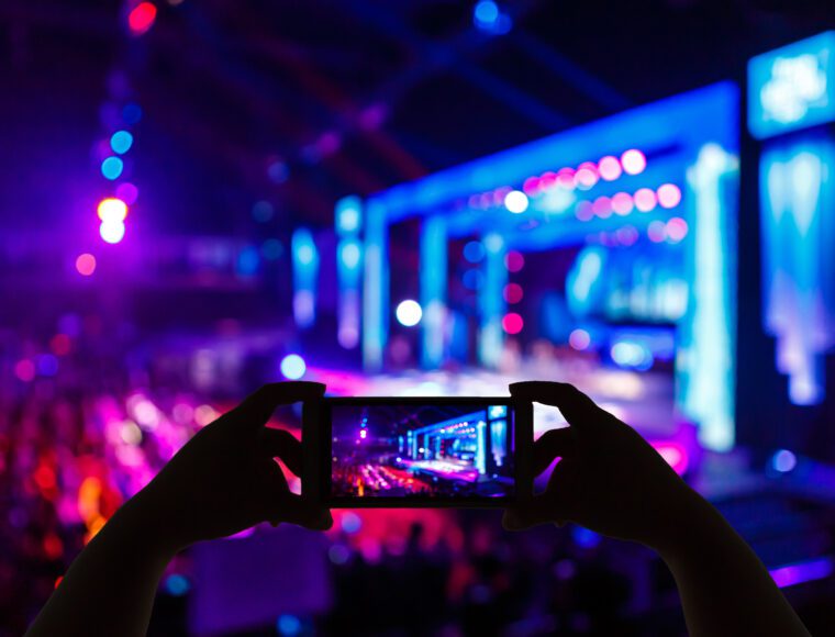 A person captures the vibrant, colorful stage at International Beatles Week on their smartphone. As bright lights dance in shades of blue, purple, and pink, the blurred stage and audience highlight the lively scene displayed on the phone's screen.
