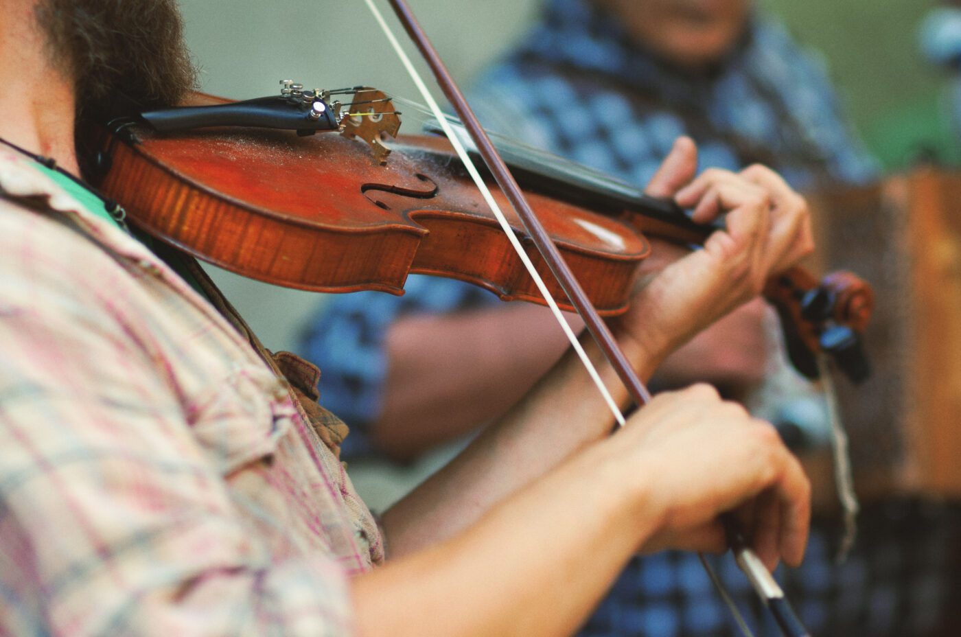 Close-up of a person playing a violin at the lively Kew Fete. The musician dons a plaid shirt, perfectly in sync with another plaid-clad individual in the background, skillfully playing an accordion.