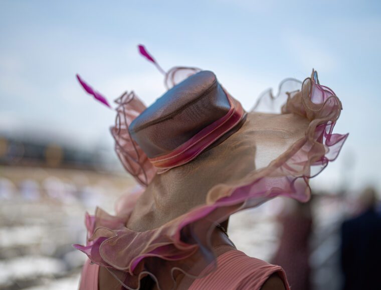 A person wearing an elaborate pastel hat adorned with pink frills and abstract feather-like decorations stands outdoors under a blue sky. The focus is on the hat's intricate design.
