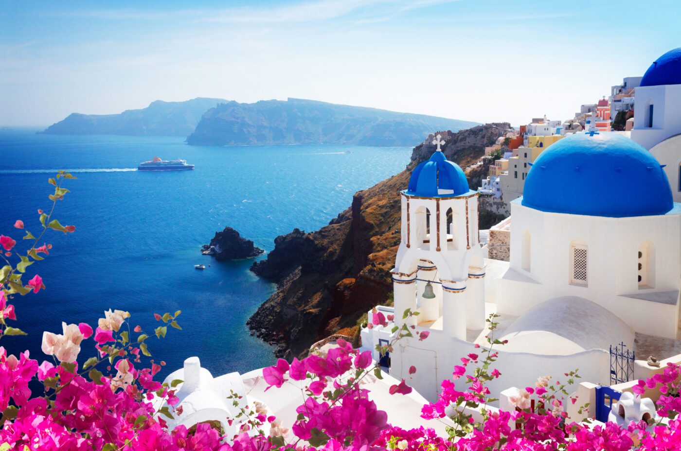 Vibrant pink bougainvillea frames white buildings with blue domes overlooking the deep blue sea in Santorini, Greece, as a cruise ship sails by. The enchanting view sets the perfect scene for an unforgettable Jazz Festival under a bright, clear sky.