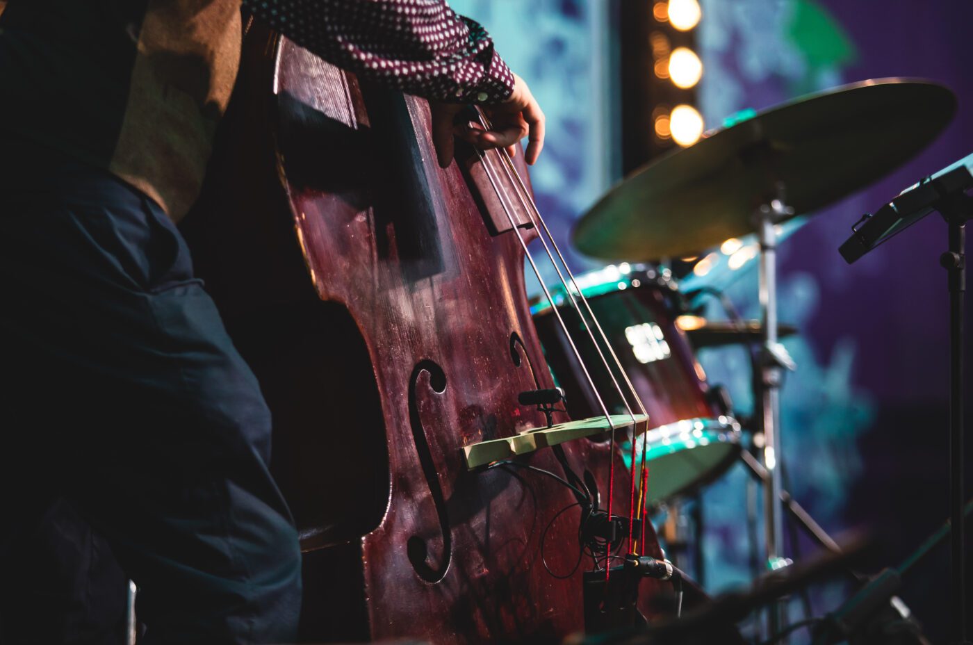 A musician plays a double bass on stage next to a drum set, with colorful lights and a blurred background creating a lively Jazz Festival atmosphere.