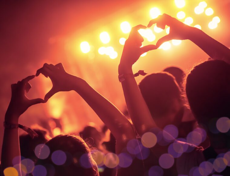A crowd at a concerts event with hands forming heart shapes silhouetted against bright, colorful stage lights. The image has a warm, vibrant glow, and out-of-focus bokeh lights enhance the festive atmosphere.