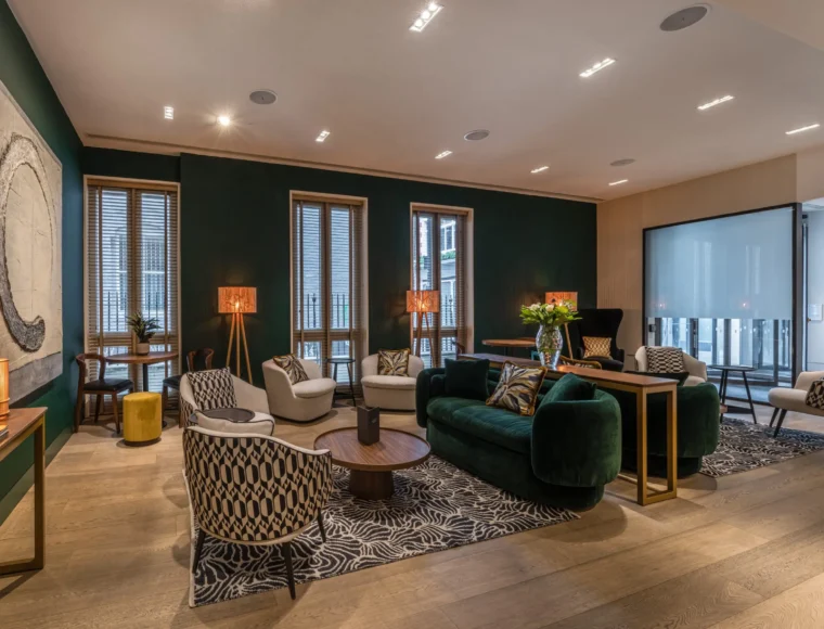 Modern living room with green walls, stylish furniture including a green velvet sofa, patterned armchairs, round wooden tables, abstract wall art, lamps, and large windows with blinds, creating a cozy and elegant atmosphere.