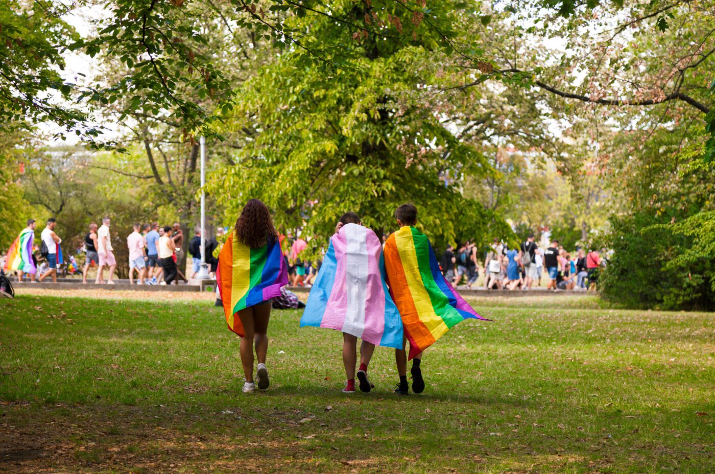 Three people walk on grass, draped in rainbow and transgender pride flags, with a crowd visible among trees—capturing the lively spirit of Edinburgh Pride