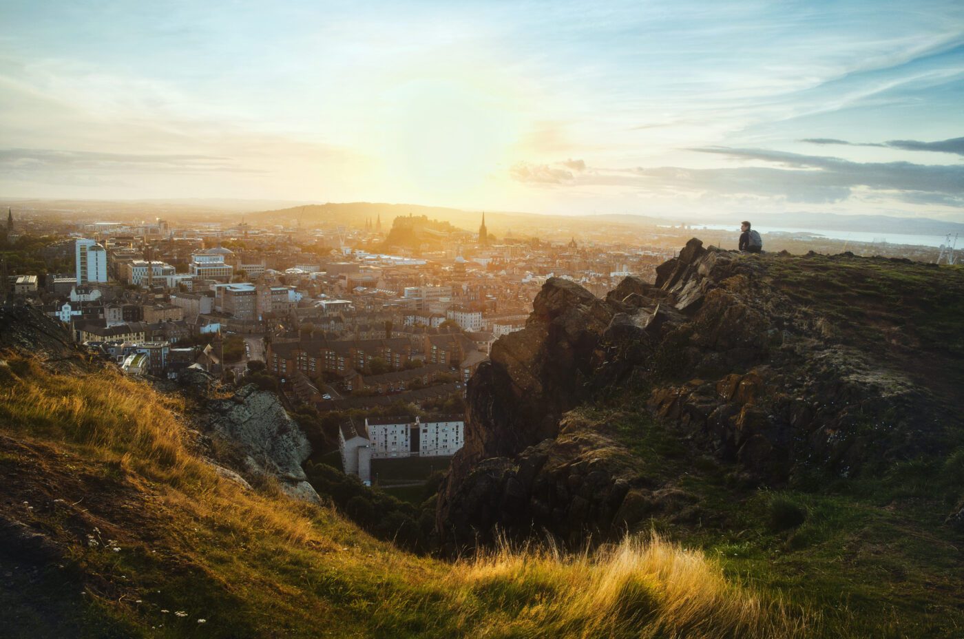 A person sits on a rocky hill overlooking a city at sunset, with sunlight illuminating buildings, distant hills, and a partly cloudy sky. Grassy slopes and rugged terrain are visible in the foreground.