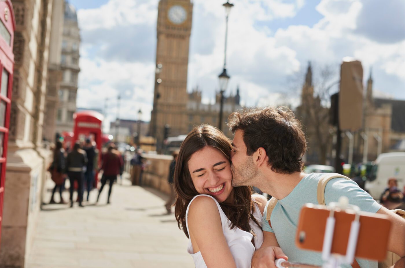 A smiling woman takes a selfie with a man who kisses her cheek on a street in London, with Big Ben and red phone booths visible in the background.