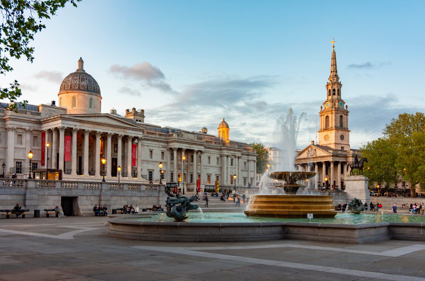 Trafalgar Square in London at sunset, showing the National Gallery on the left, the church of St Martin-in-the-Fields on the right, and a central fountain with people sitting around the square.