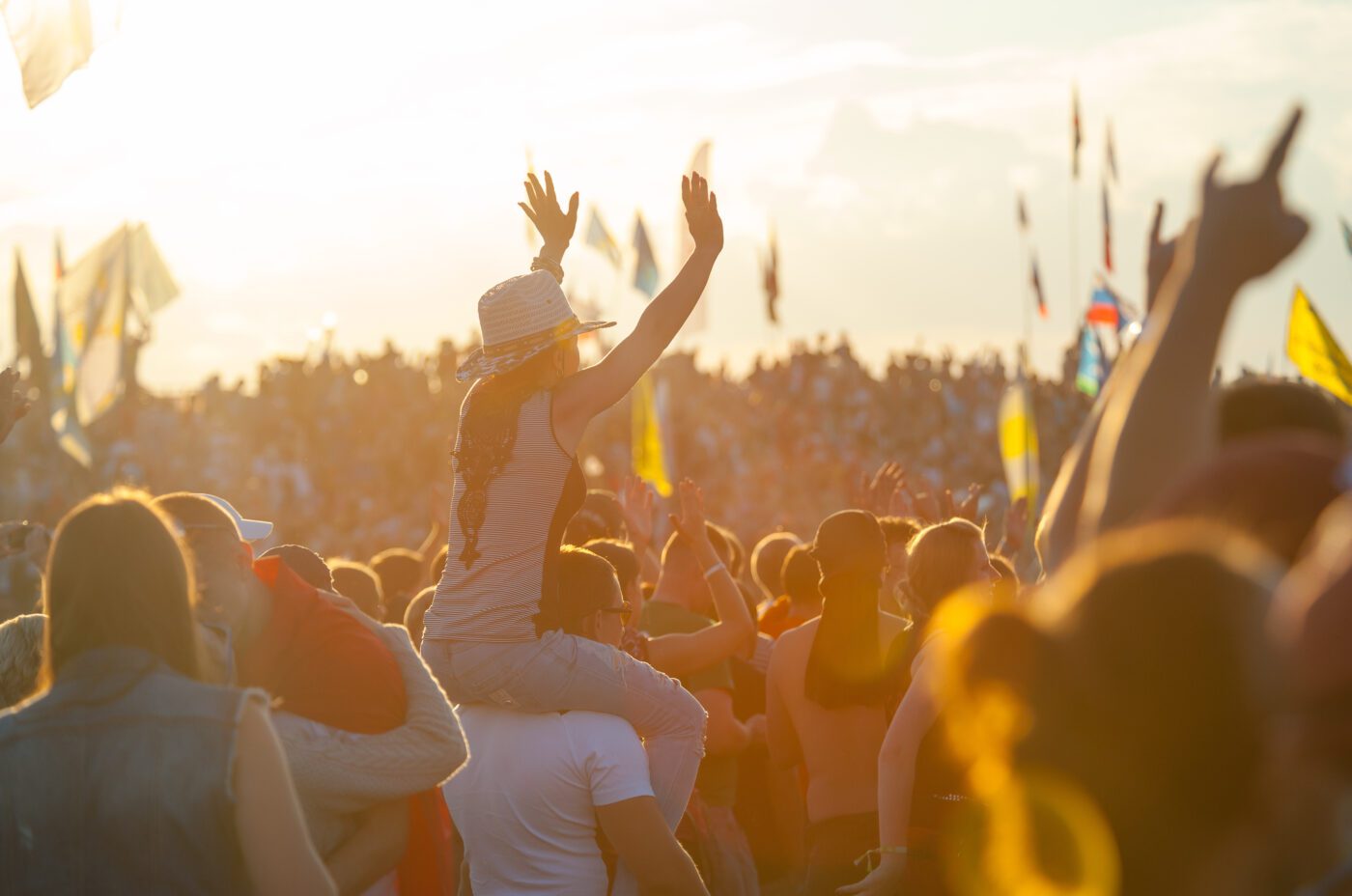 A crowd enjoys an outdoor festival during sunset. Amidst the energy of International Beatles Week, a person in a hat sits on someone’s shoulders, arms raised high, as flags wave and music fills the bright, lively scene.