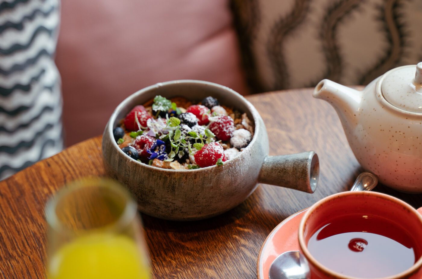 A bowl of granola with fruit and seeds, a cup of tea with a Dishoom teapot, and a glass of orange juice on a wooden table, with patterned pillows in the background.