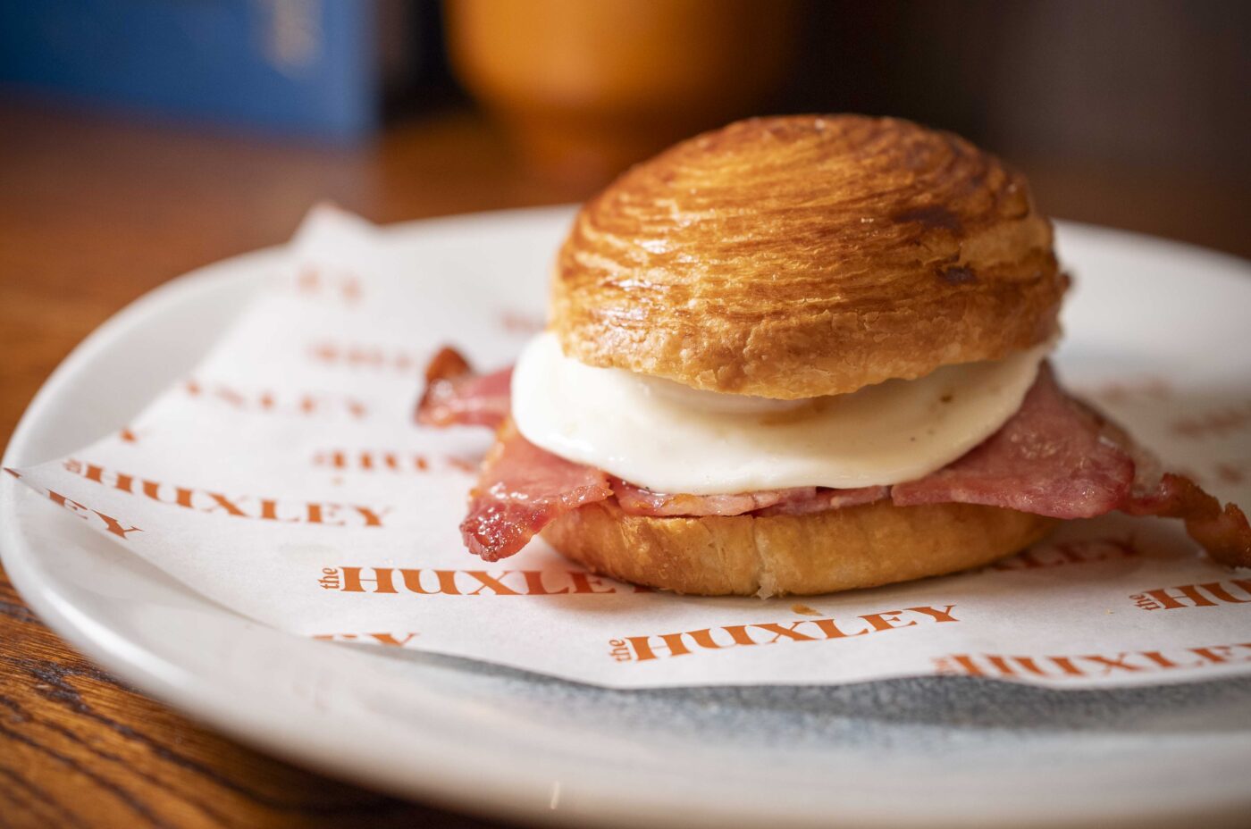 A croissant sandwich with ham and melted cheese rests on branded paper atop a white plate on a wooden table, with Dishoom’s signature style evident amid a blurred plant and book in the background.