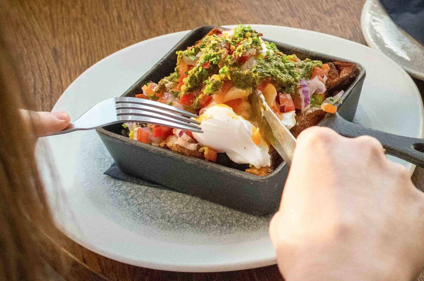 A person uses a fork and knife to cut into a Dishoom-inspired dish topped with a poached egg, diced vegetables, and green sauce, served in a small rectangular skillet on a white plate.