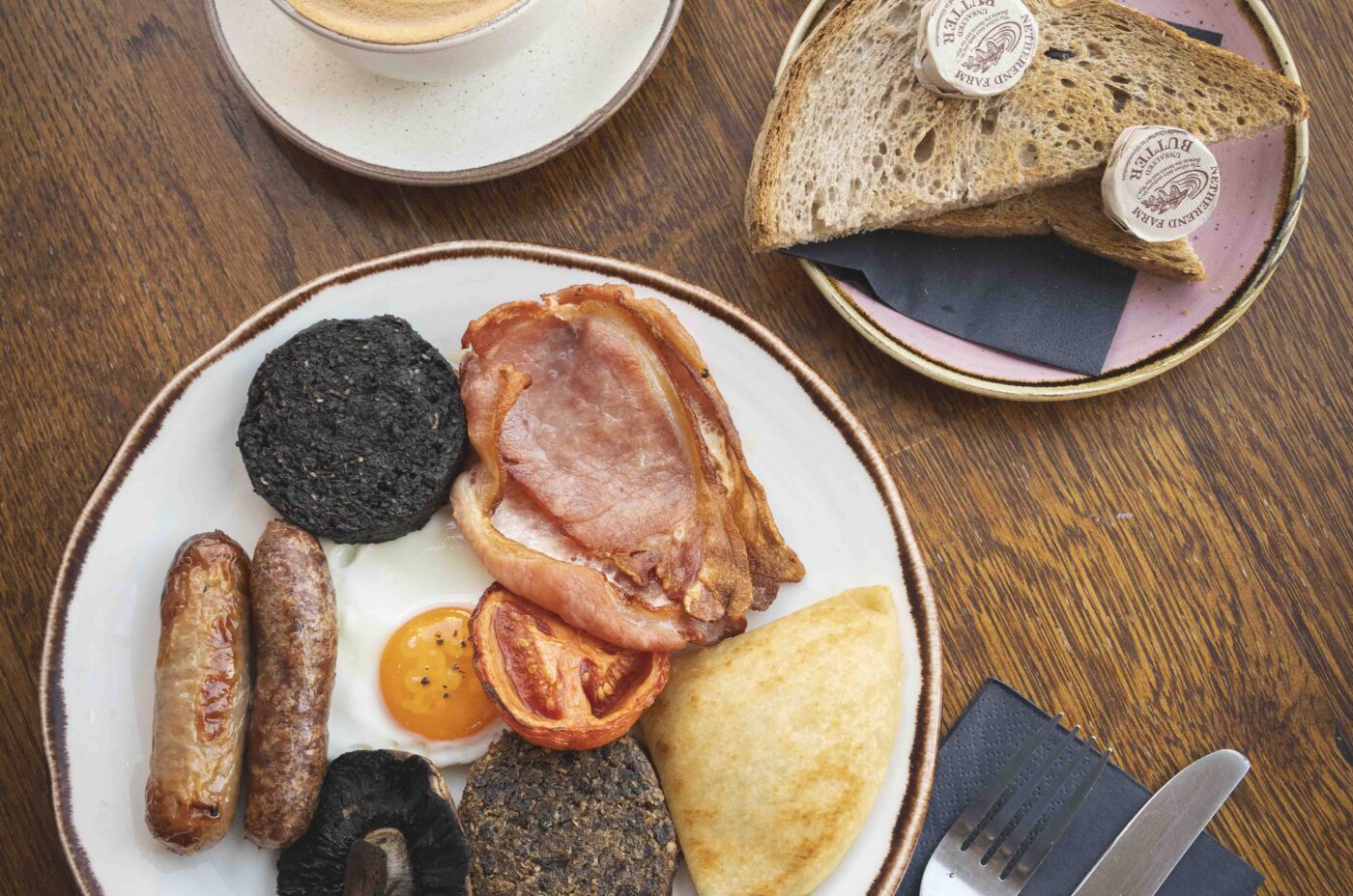 A full breakfast plate with bacon, sausages, black pudding, egg, grilled tomato, mushroom, and potato bread sits beside a cup of latte with latte art—a Dishoom-inspired spread—with buttered wholemeal toast and cutlery on a napkin.