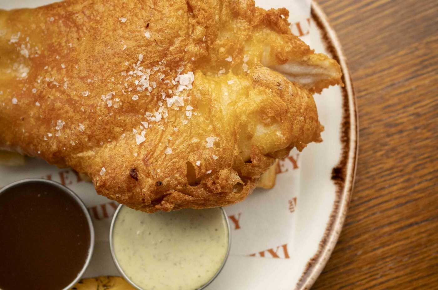 A plate with a large piece of battered fried fish, a wedge of grilled lemon, two dipping sauces, and mushy peas on parchment paper, set on a wooden table—a Dishoom-inspired treat.