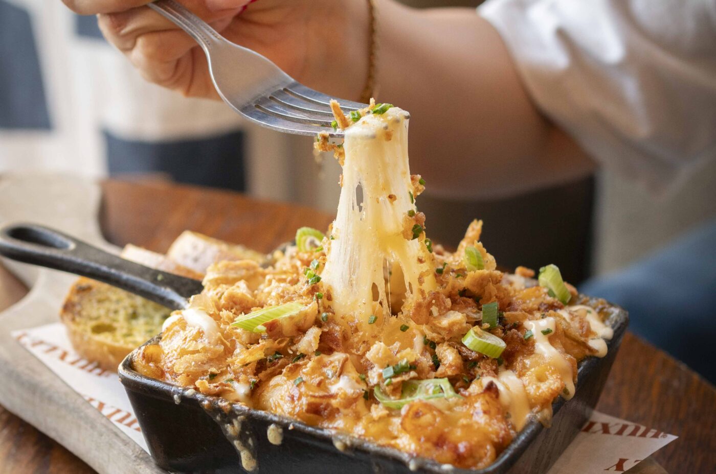 A hand lifts a forkful of cheesy baked pasta from a small skillet, with melted cheese stretching, green onions and breadcrumbs on top—a Dishoom-inspired delight—served on a wooden board with toasted bread on the side.