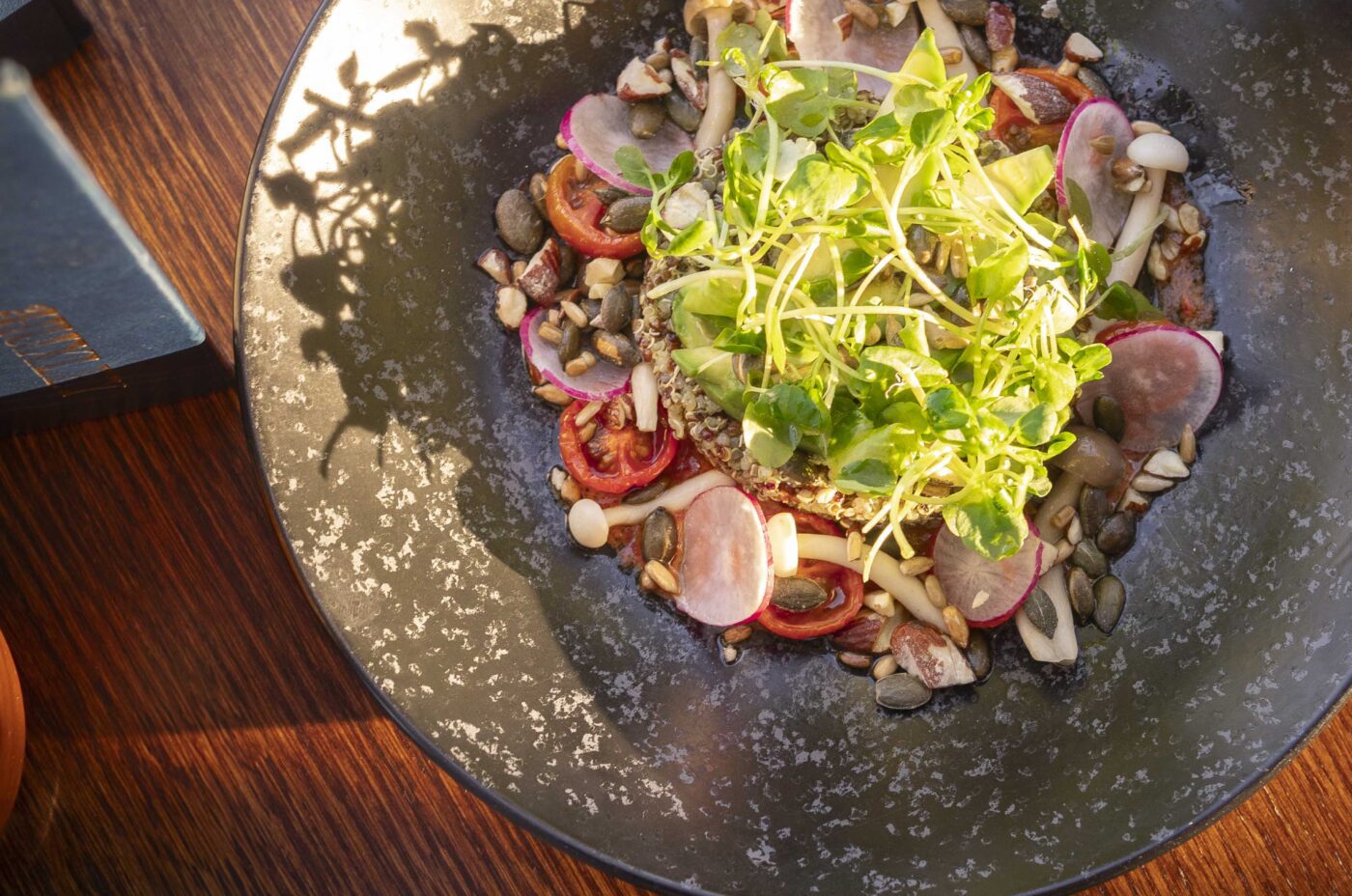 A black speckled bowl from Dishoom holds a vibrant salad of microgreens, radish slices, cherry tomatoes, mushrooms, and seeds, sitting on a wooden table bathed in natural sunlight.