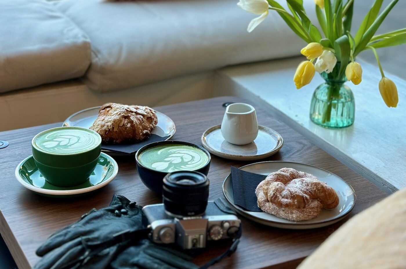 A cozy Bon Vivant café scene with two matcha lattes, pastries, a camera, black gloves, and a vase of yellow tulips on a wooden table by a window seat with striped pillows and a street view outside.