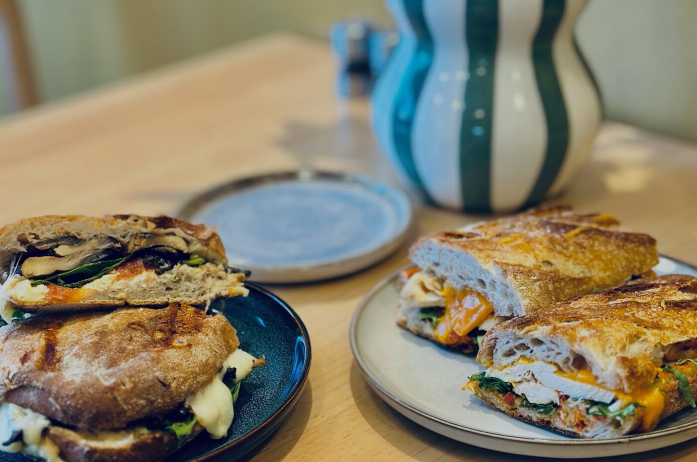 Two plates of toasted sandwiches filled with vegetables, cheese, and greens sit on a wooden table—perfect for any Bon Vivant. A striped ceramic vase and an empty plate are in the background.