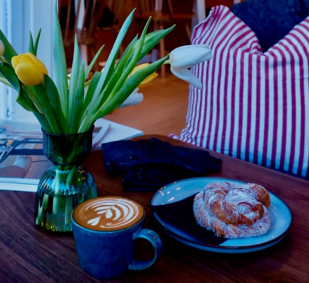 A cozy Bon Vivant cafe scene with a cup of latte, a pastry on a plate, a vase of yellow tulips, and a striped pillow on a chair. Wooden chairs and tables with flowers in the background add to the warm atmosphere.