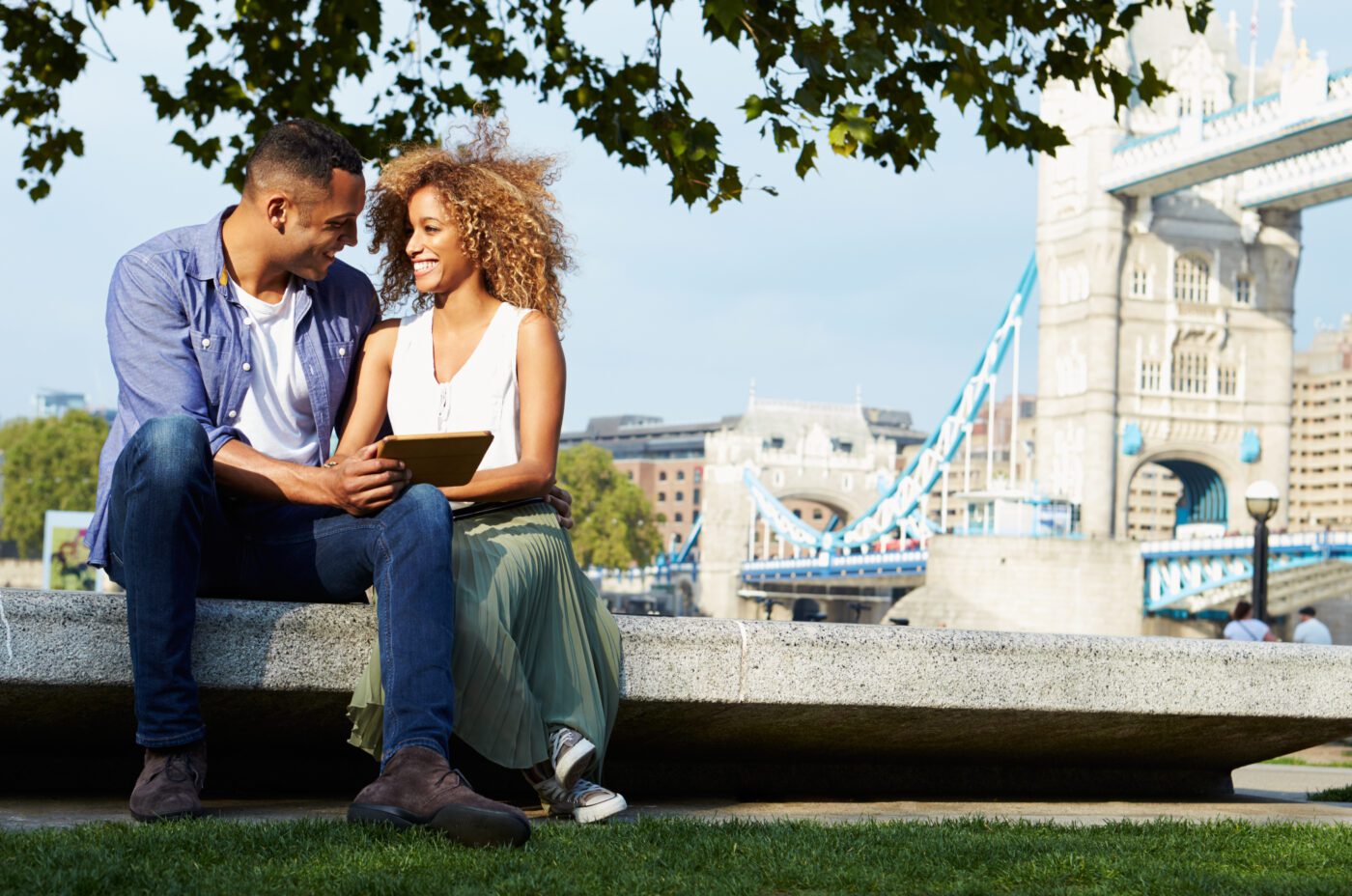 A smiling couple sits on a stone bench in a park, looking at a tablet together, with the iconic Tower Bridge in London visible in the background under a partly sunny sky.