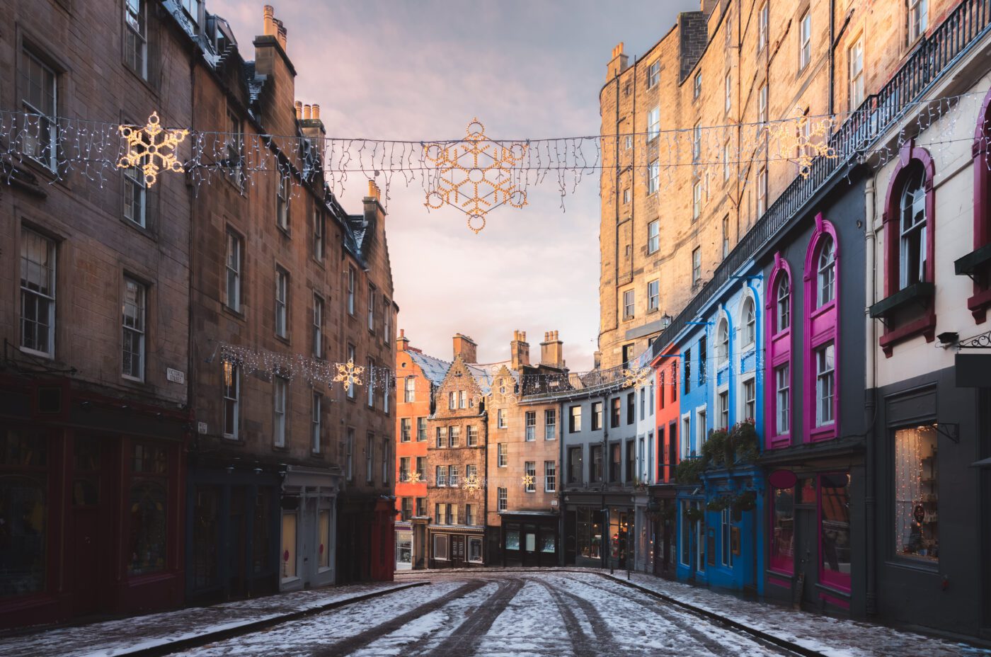 A quiet, snow-dusted street lined with tall, historic buildings—some with colorful pink and blue facades—hosts festive EDI banners and snowflake ornaments strung overhead beneath a soft, cloudy sky.