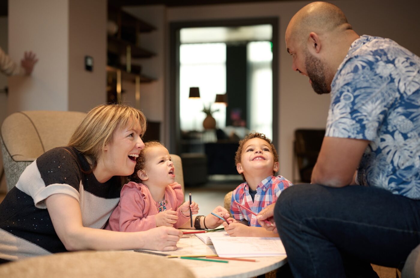 A smiling family of four sits around a table, coloring and laughing together in a cozy living room. The parents engage warmly with their two young children, creating a joyful and playful atmosphere.