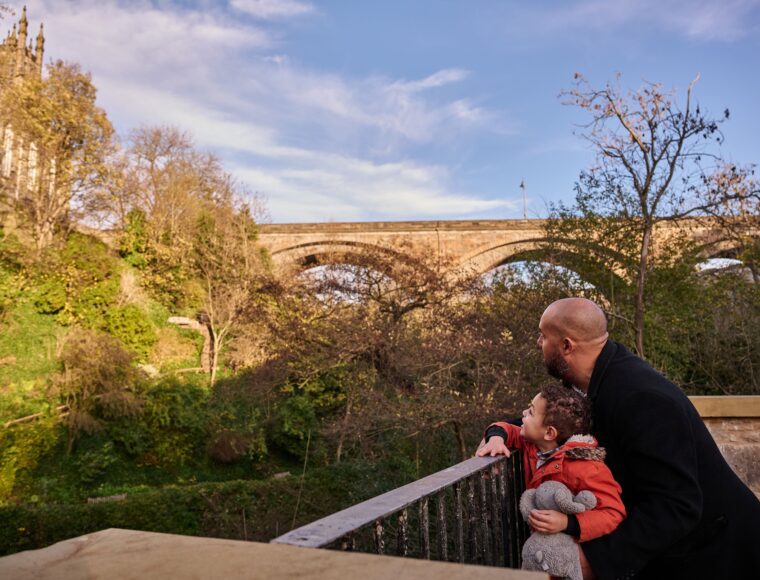A man and a child in warm clothes look up at an old stone bridge surrounded by trees and greenery on a sunny day.