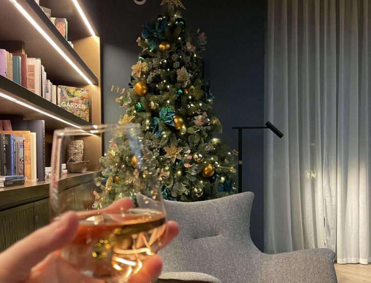 A hand holds a glass of wine in a cozy room with edi-inspired decor, a decorated Christmas tree, bookshelves, a modern armchair, and soft lighting.
