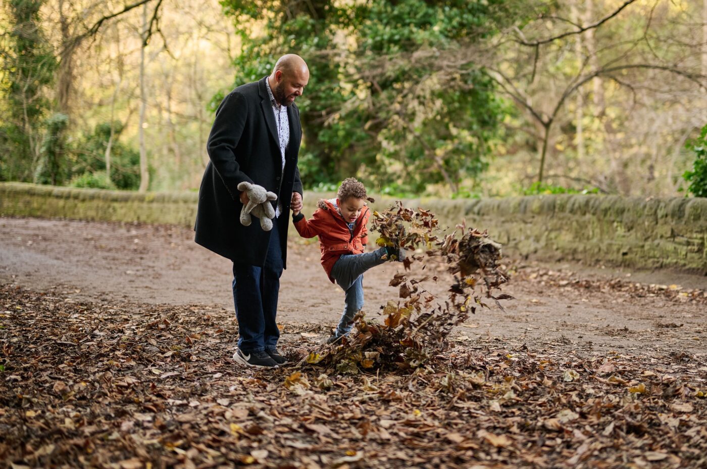 A man holding a stuffed animal and a child’s hand walks outdoors. The child, wearing a red jacket, is kicking up a pile of fallen leaves on a path surrounded by trees.