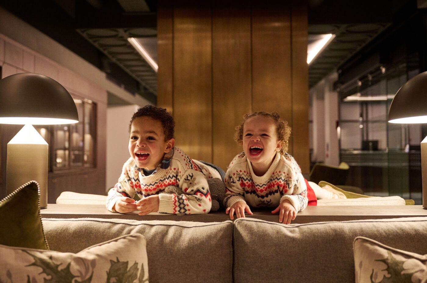 Two young children wearing matching festive sweaters lie on their stomachs on the back of a sofa, smiling and laughing brightly in a warmly lit, modern edi-inspired room.