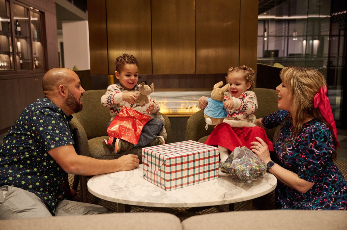 A family of four sits around a table with a gift box. Two young children, smiling, hold stuffed bunnies, while the adults look on. Amidst unwrapped paper and a cozy indoor setting, they share an edi moment together.