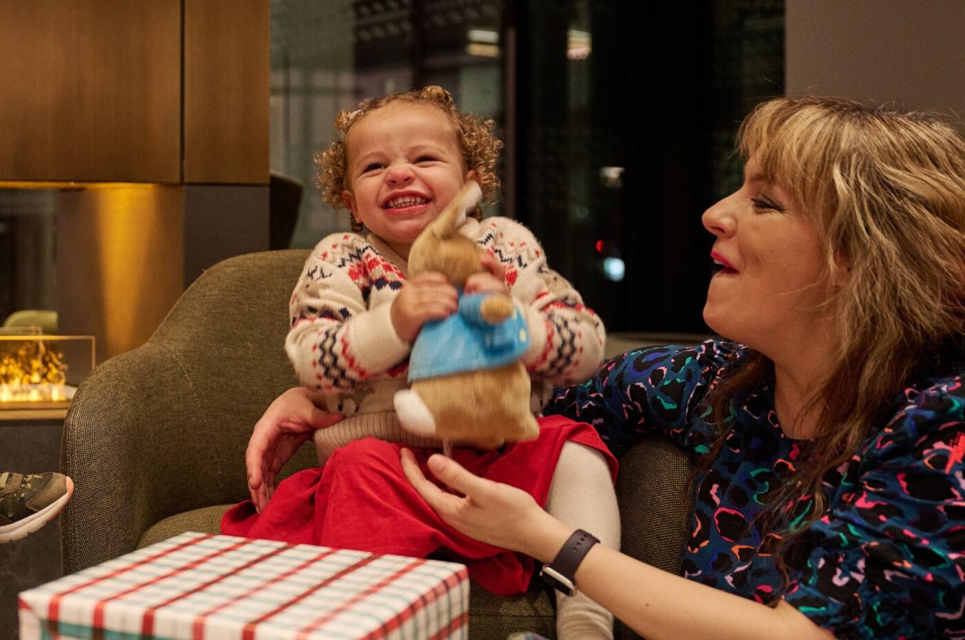 A young child smiles joyfully while holding a stuffed bunny, sitting on a couch beside an adult woman. Wrapped gifts are on the table in front of them, and the warmth of the glowing fireplace adds to the cozy edi atmosphere.