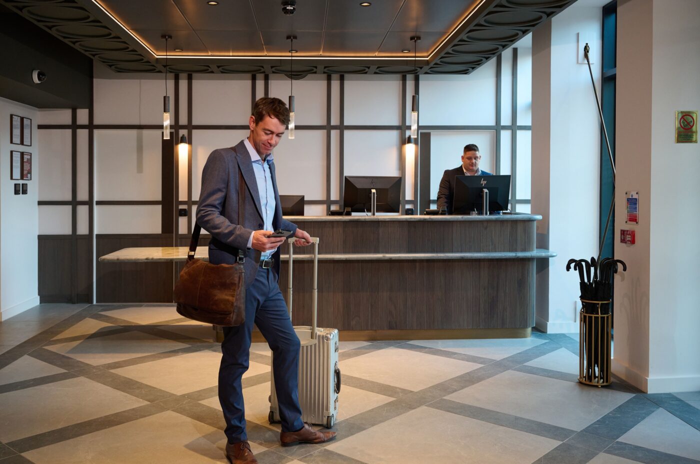 A man in a suit stands in a hotel lobby, looking at his phone with a suitcase and shoulder bag. A receptionist behind the counter smiles at him. The lobby is modern and well-lit.