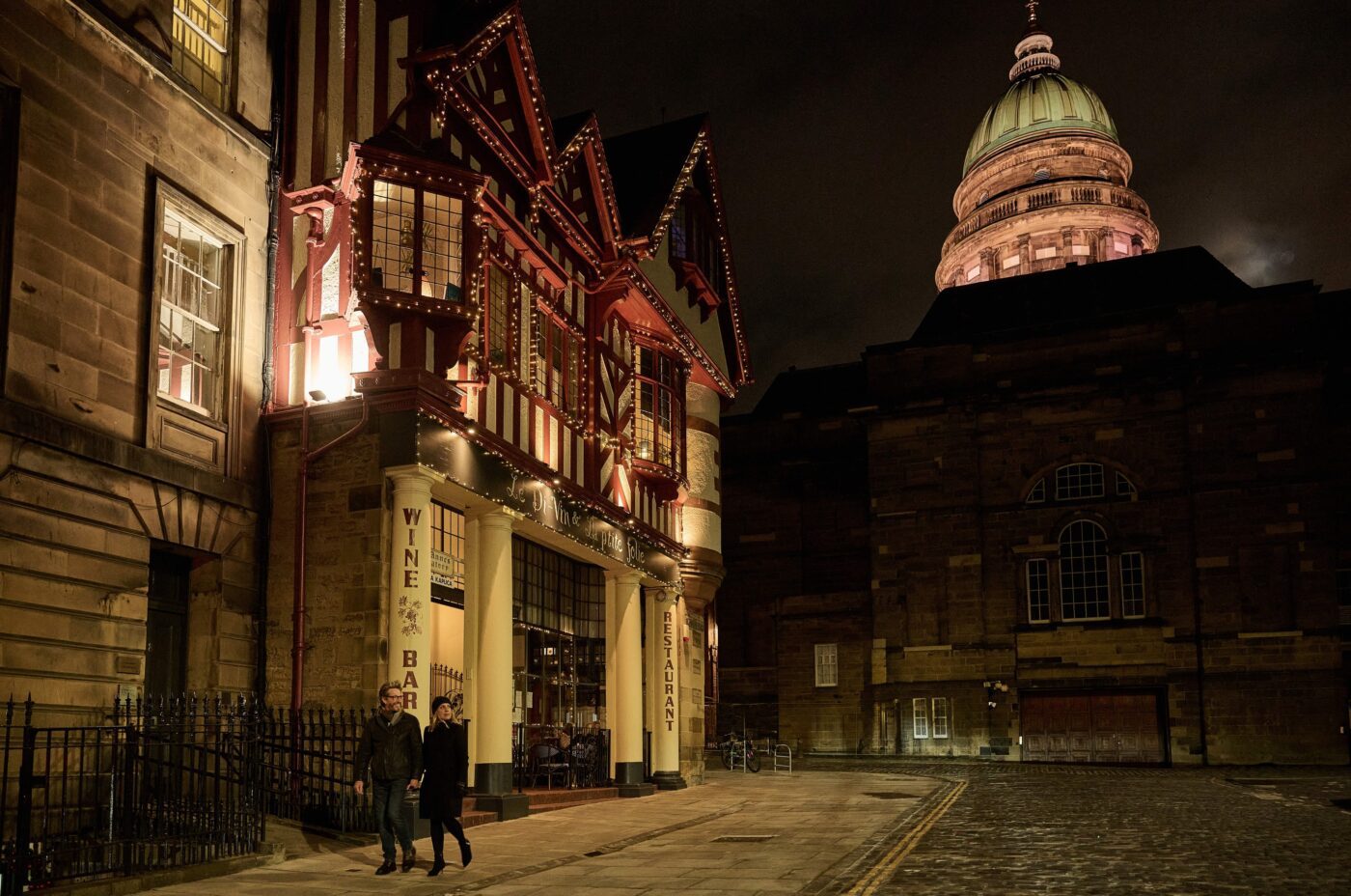 Two people walk past a warmly lit wine bar on a cobblestone street at night, with the historic edi building featuring a domed roof illuminated in the background.