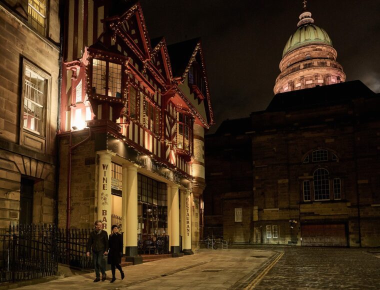 Two people walk past a warmly lit wine bar on a cobblestone street at night, with the historic edi building featuring a domed roof illuminated in the background.
