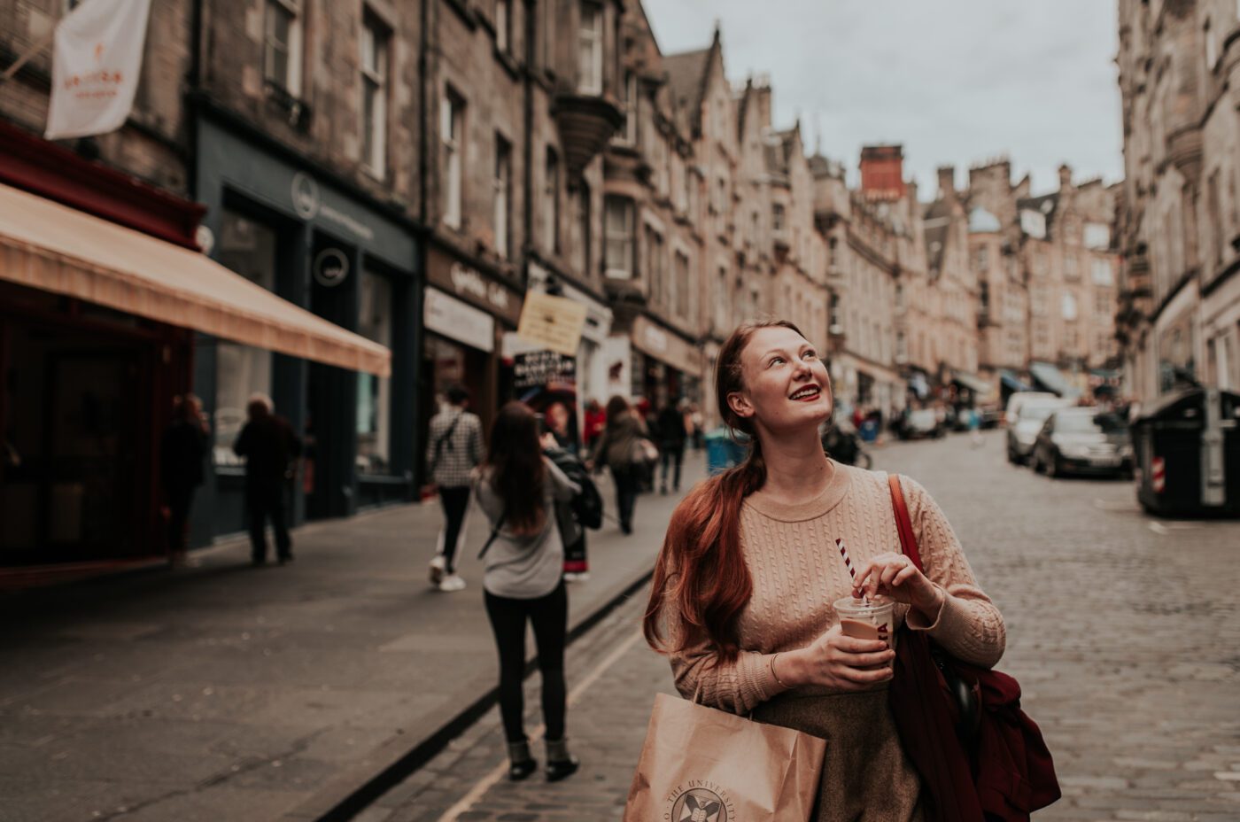 A young woman with red hair smiles and looks up while holding a drink and shopping bag on a cobblestone street lined with historic buildings and people walking.