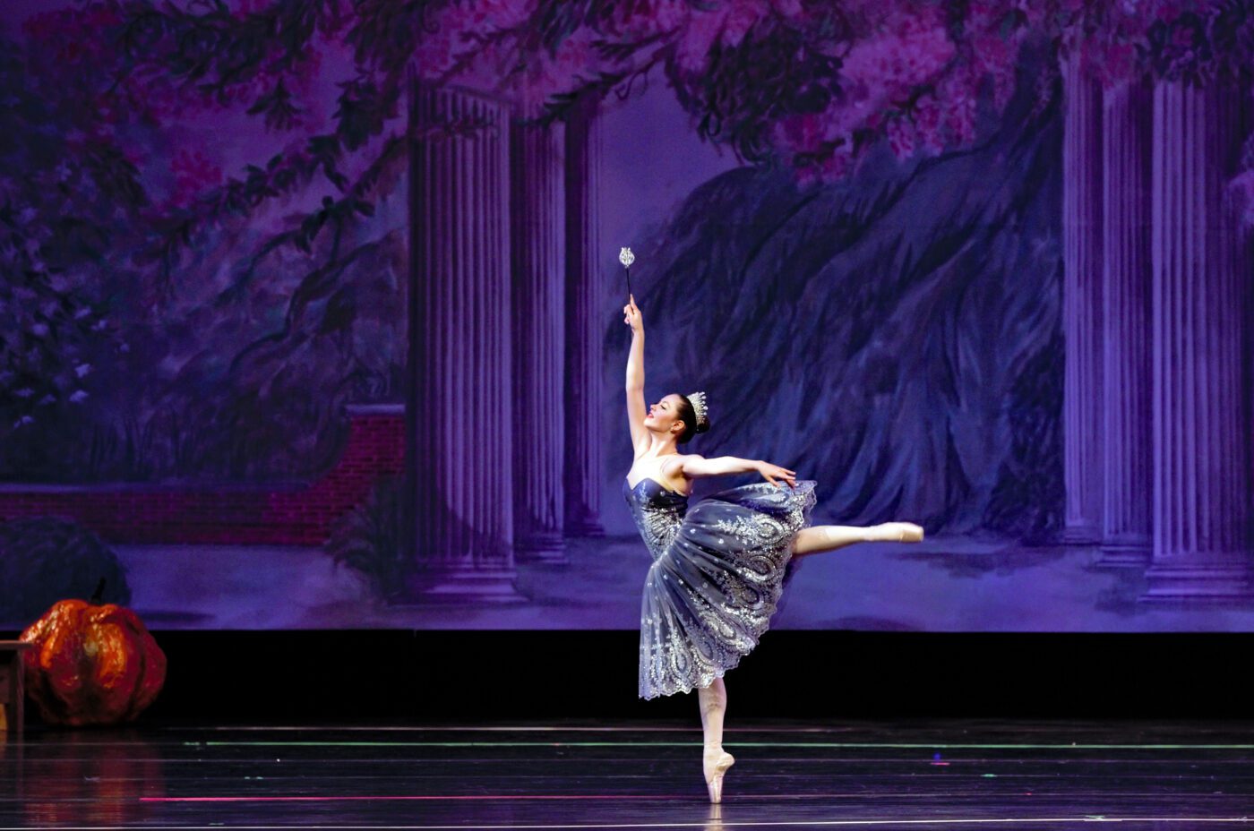 A ballerina in a sparkling dress and tiara dances on stage, standing en pointe with one leg extended behind her and one arm raised, evoking a wicked grace against a backdrop of classical columns and purple foliage.