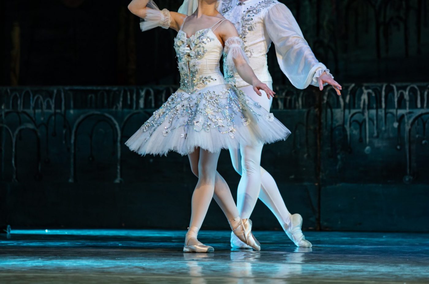 Two ballet dancers in white, ornate costumes perform at Kew Fete. The female dancer wears a tutu and pointe shoes, while the male stands behind her, both poised gracefully against a dark, ornate backdrop.