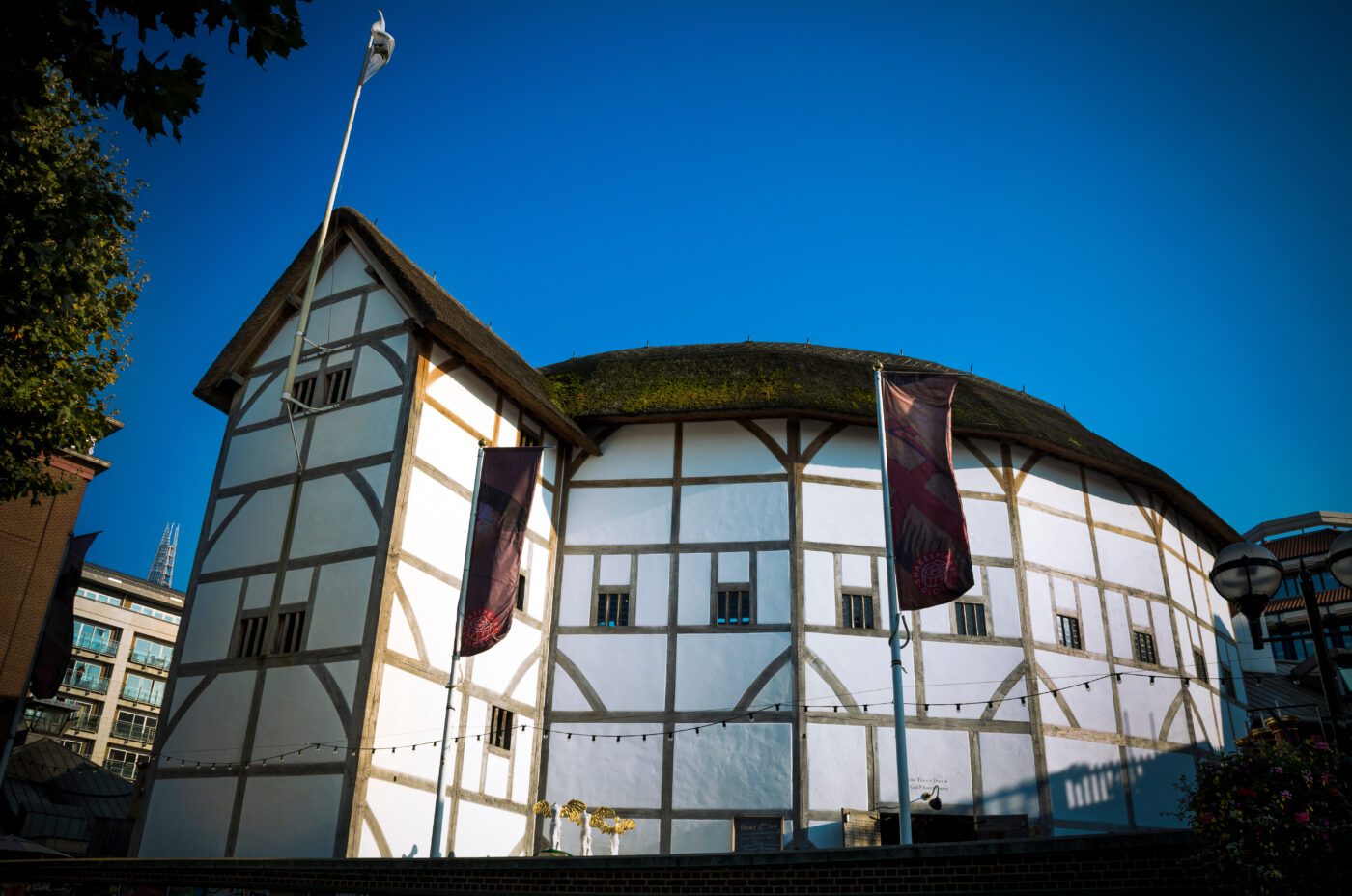 A white, timber-framed circular building with a thatched roof stands under a clear blue sky at the Kew Fete; two vertical banners hang by the entrance, and a flag flies from the roof.