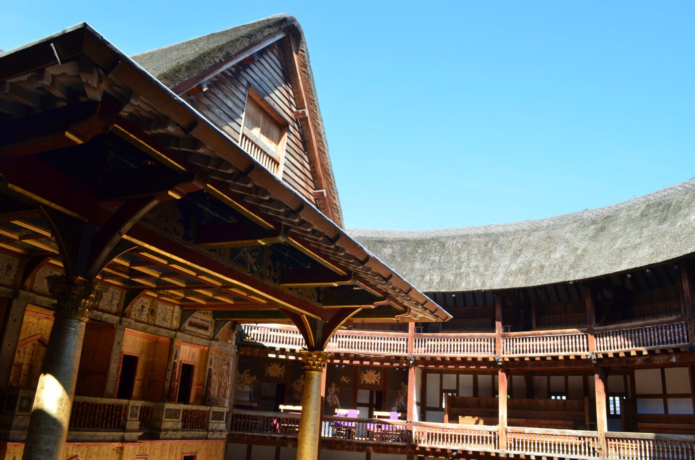 Open-air view of a wooden, circular theater with thatched roof and multiple balconies, featuring ornate columns and detailed architecture—reminiscent of the lively atmosphere found at Kew Fete—under a clear blue sky.