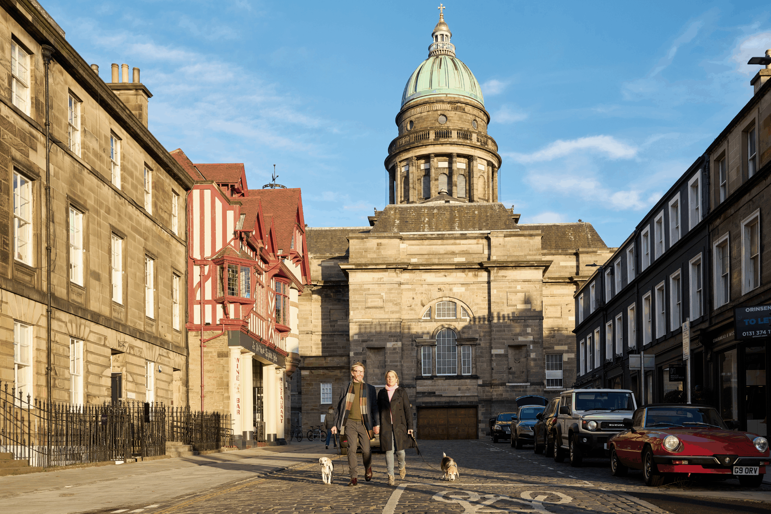 Two people walk dogs down a cobblestone street lined with historic buildings, with a domed stone building in the background under a clear blue sky. Several parked cars are visible on the right side of the street.