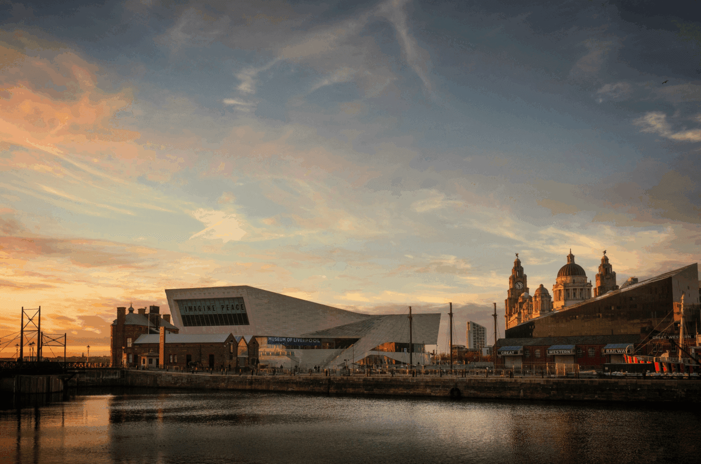 A waterfront cityscape at sunset shows modern and historic buildings, including the angular Museum of Liverpool, with warm light reflecting on the water and a dramatic, cloud-filled sky overhead.