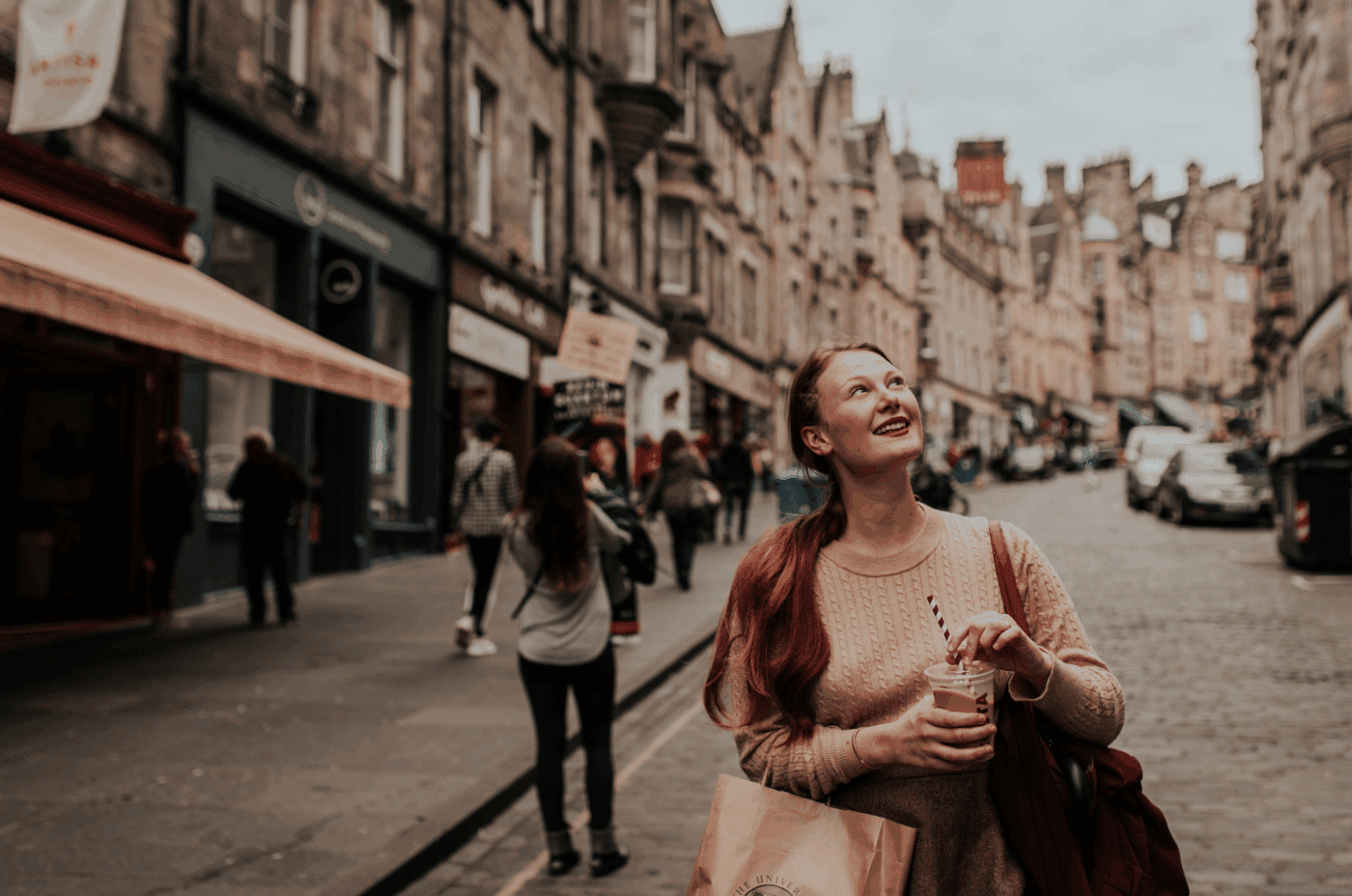 A woman with long red hair smiles and looks up while holding a drink on a cobblestone city street lined with historic buildings and shops. Other people walk in the background.