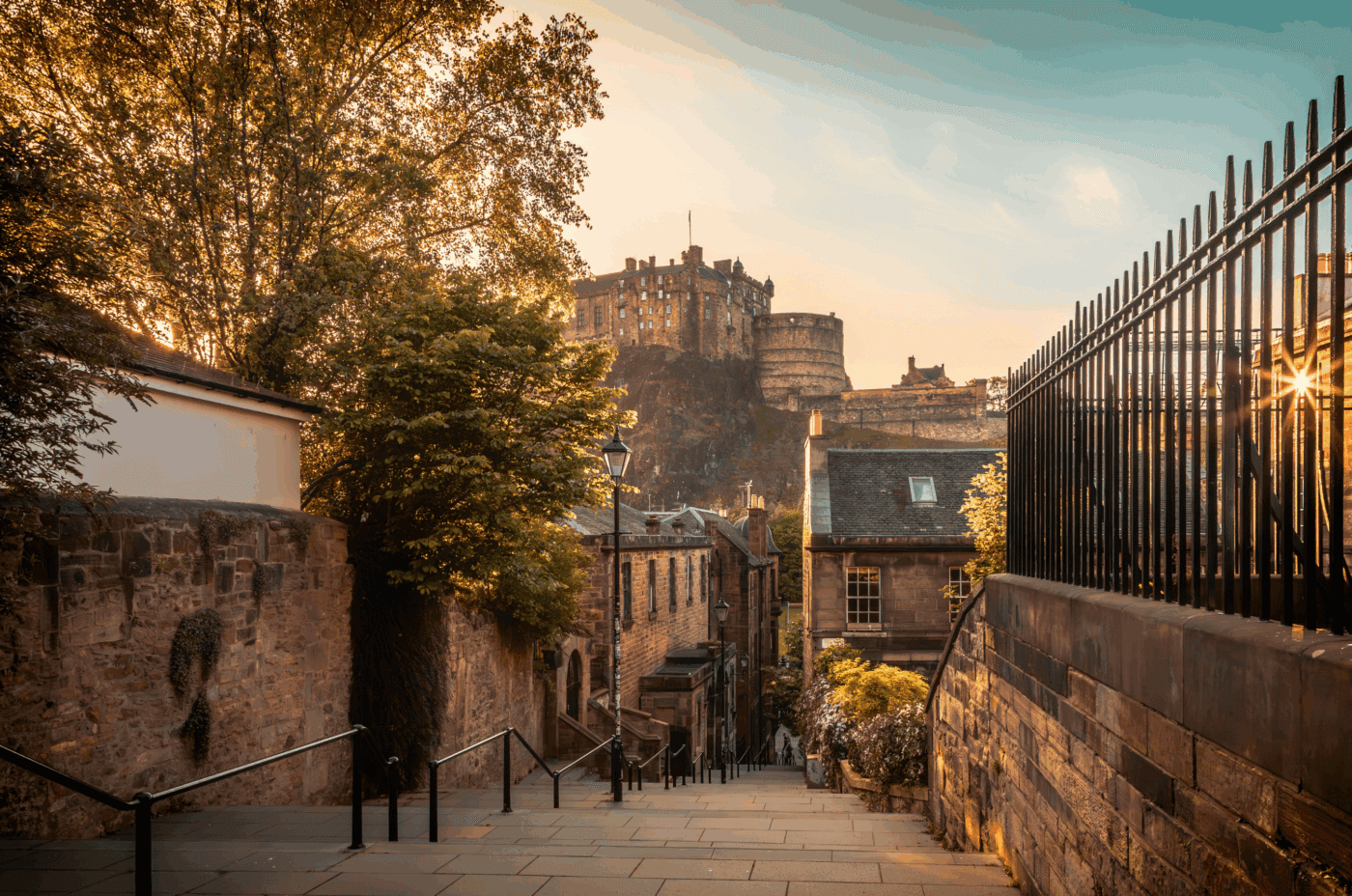 A sunlit, cobblestone street lined with stone buildings and greenery leads up to a historic castle perched on a rocky hill under a partly cloudy sky.