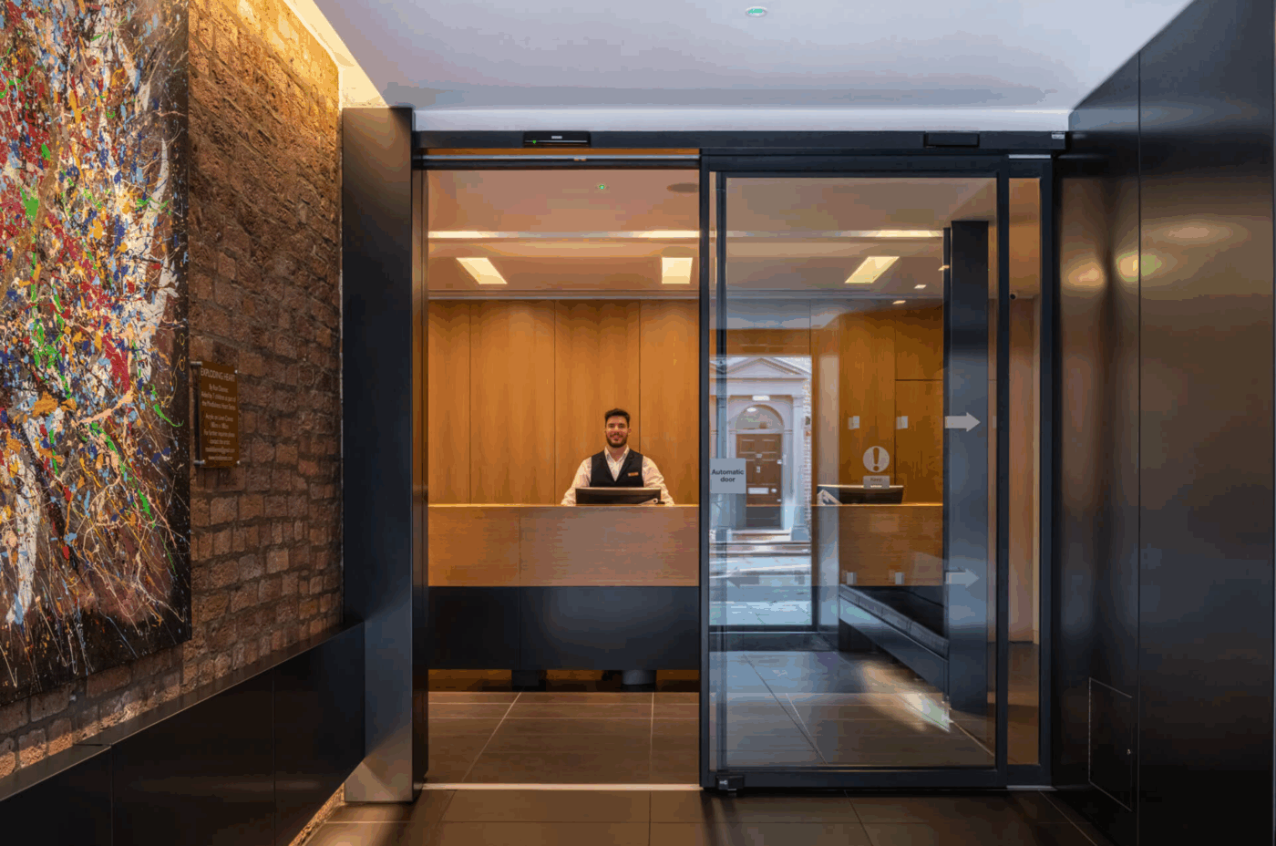 A receptionist stands behind a wooden desk in a modern, well-lit lobby with glass doors. The walls are dark with one featuring a large, colorful abstract painting.