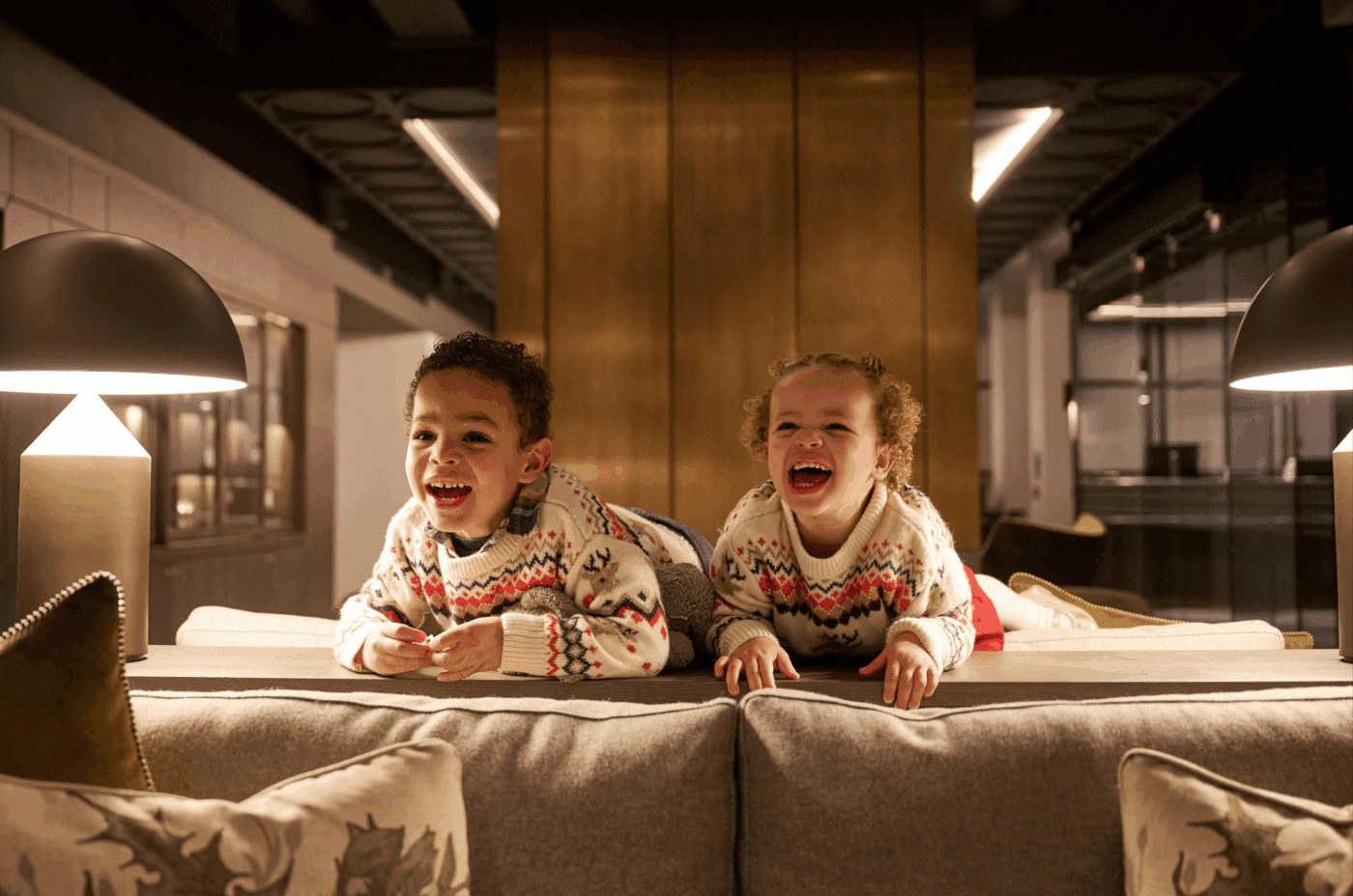 Two young children wearing matching festive sweaters lie on their stomachs on the back of a sofa, laughing and smiling under warm indoor lighting between two table lamps.