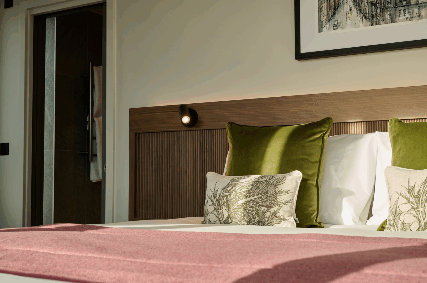 A cozy bed with green and botanical pillows, white sheets, a pink blanket, a wooden headboard, and a wall-mounted reading light, with sunlight streaming in from the left and an open door visible in the background.