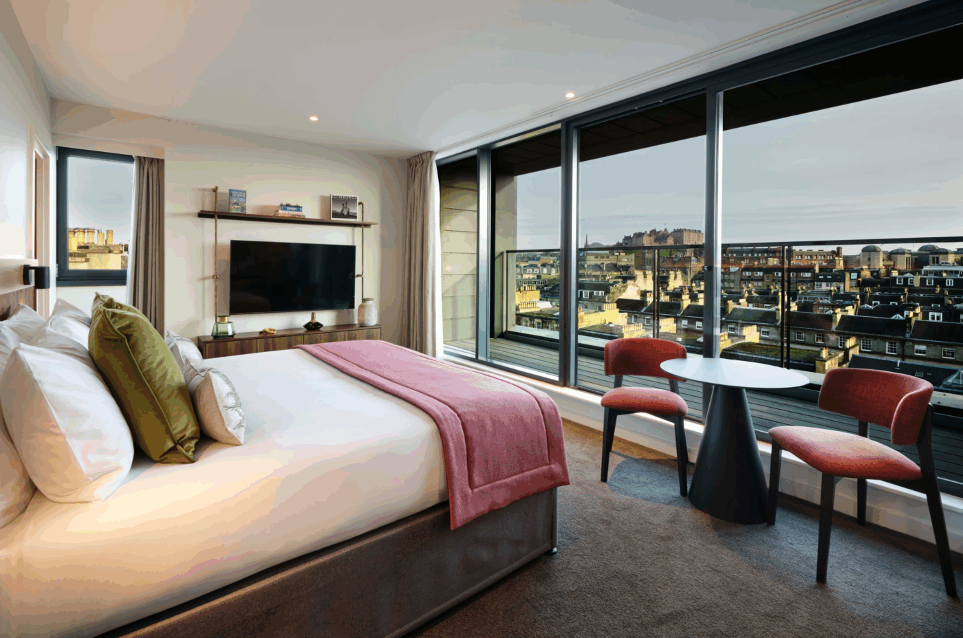 A modern hotel room with a large bed, TV, two red chairs with a small black table, and floor-to-ceiling windows offering a cityscape view at dusk.