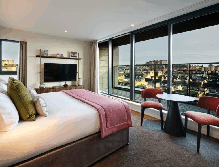 A modern hotel room with a large bed, TV, two red chairs with a small black table, and floor-to-ceiling windows offering a cityscape view at dusk.