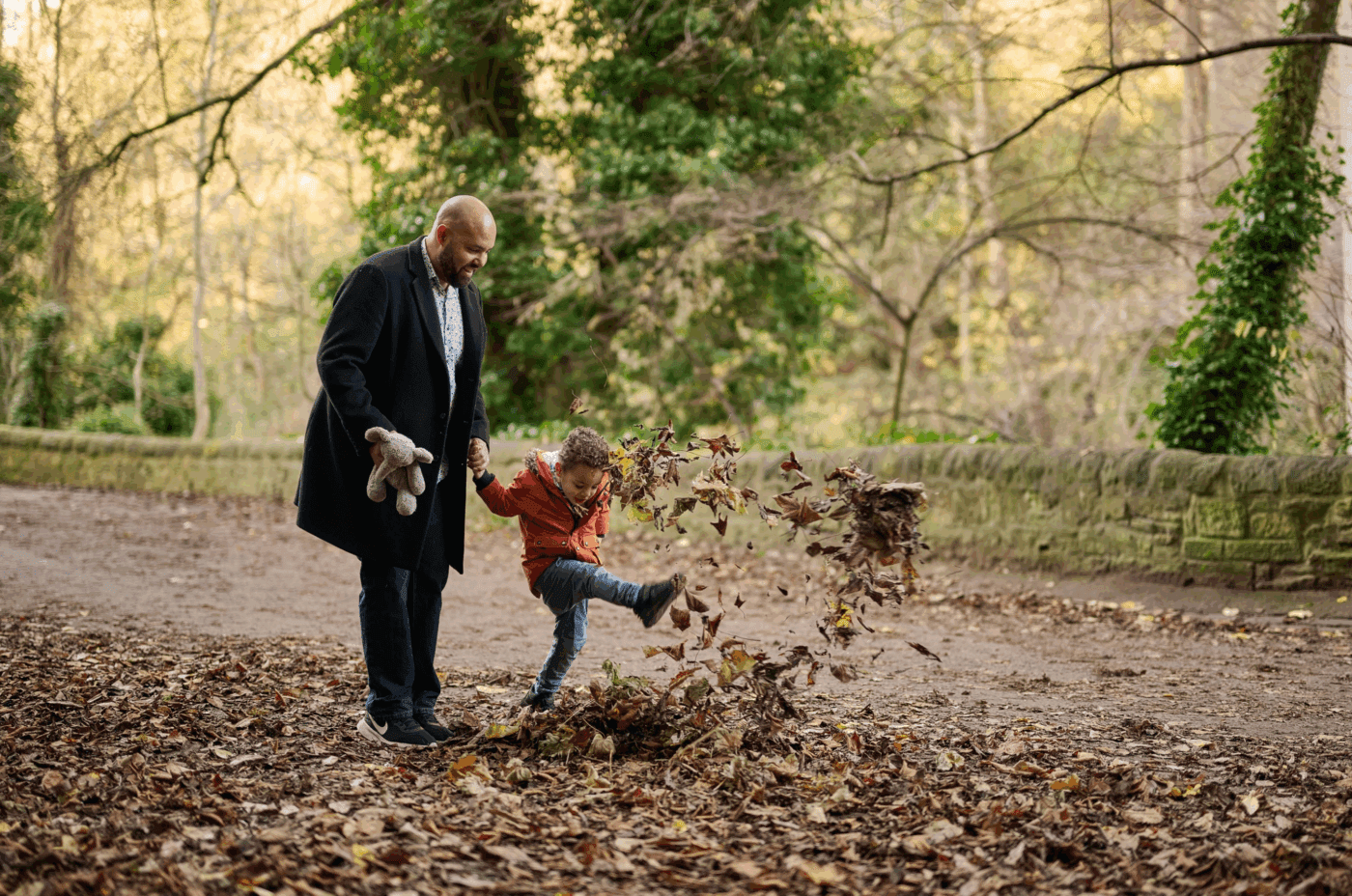 A man holding a stuffed animal stands beside a young boy who is joyfully kicking a pile of fallen leaves on a tree-lined path in a park.