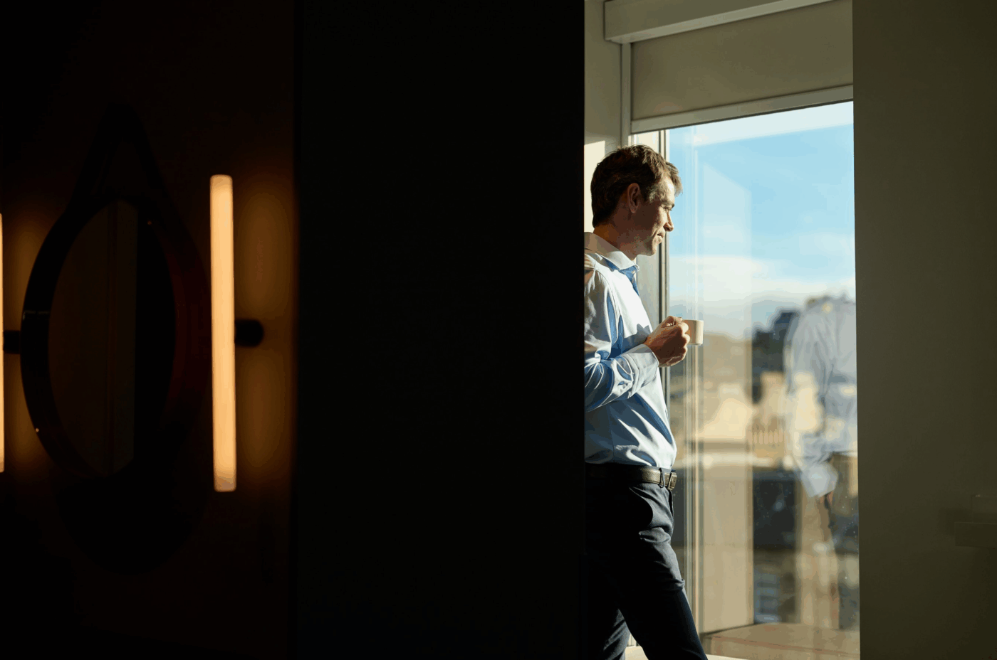 A man in a blue shirt stands by a bright window, holding a cup and looking outside thoughtfully. The room around him is dimly lit, creating a contrast with the sunlight streaming in.